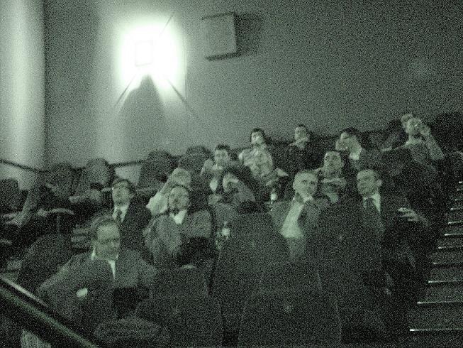 Grupa mężczyzn w garniturach siedzi na widowni podczas szkolenia lub konferencji, oświetlona przez kwadratowe okno z tyłu sali.